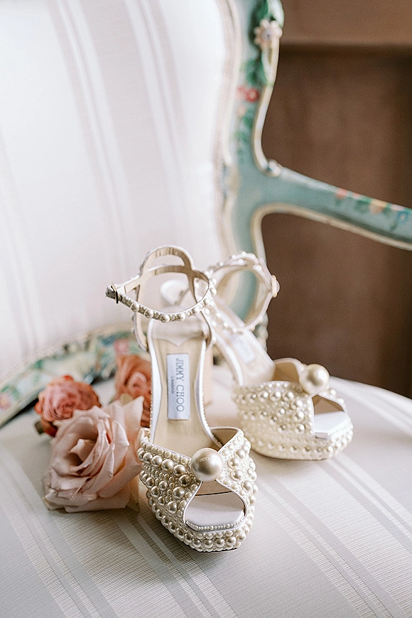 Bridal shoes, pearl wedding heels with ankle straps and satin finish resting on an upholstered chair beside roses on striped fabric surface