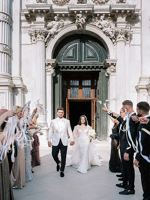 Wedding exit as newlyweds walk down ornate church steps, bride in lace dress and veil holding bouquet while guests wave ribbon wands