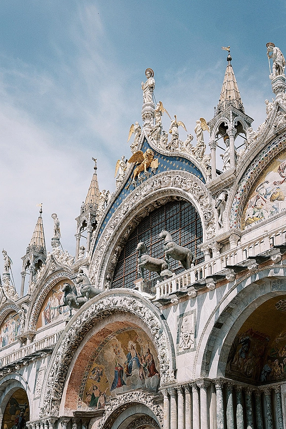 Cathedral exterior with ornate church facade, featuring spires, arches, statues, mosaic panels, and a gilded winged lion under blue sky