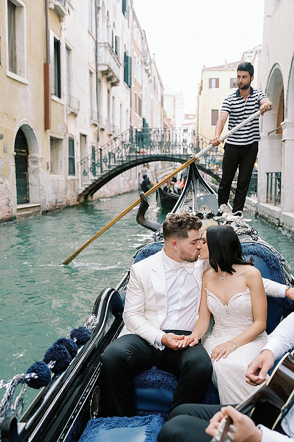 Wedding kiss as newlyweds on gondola embrace in a strapless gown and tuxedo, drifting past canal water, arched bridge, and historic buildings