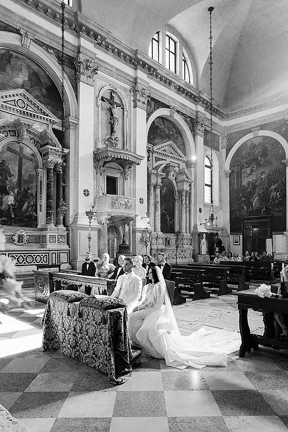Ceremony moment as bride and groom kneel at the altar with her long veil and train, framed by an ornate church interior with arched windows