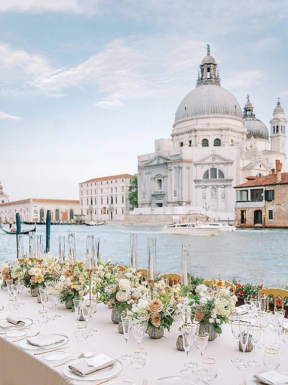 Reception tablescape with blush and white rose centerpieces, greenery, and cylinder candles on a long table by a canal with boats.