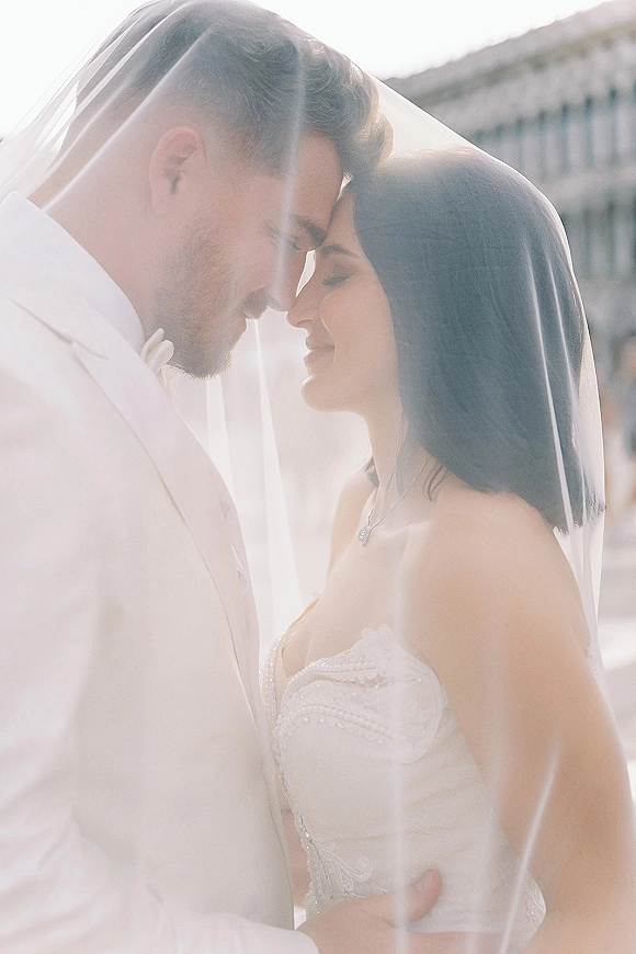 Wedding couple portrait with bride and groom forehead touch under a bridal veil, groom in white suit, daylight building exterior behind