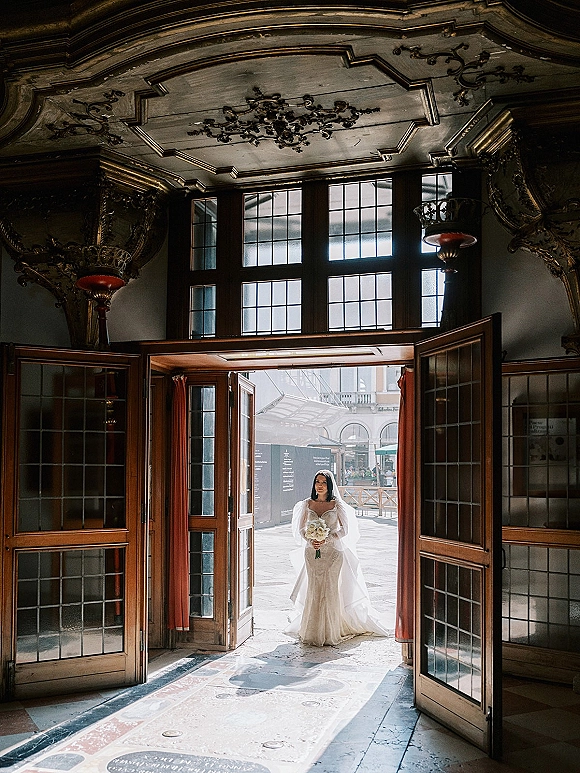 Bridal portrait of a bride holding bouquet in a lace wedding dress and veil, framed by grand wooden doors in an ornate entry hall