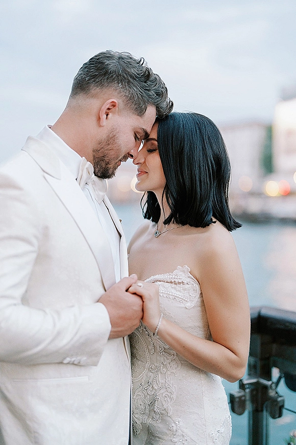 Couple portrait of bride and groom portrait touching foreheads and holding hands, bride in wedding dress by waterfront railing with city skyline