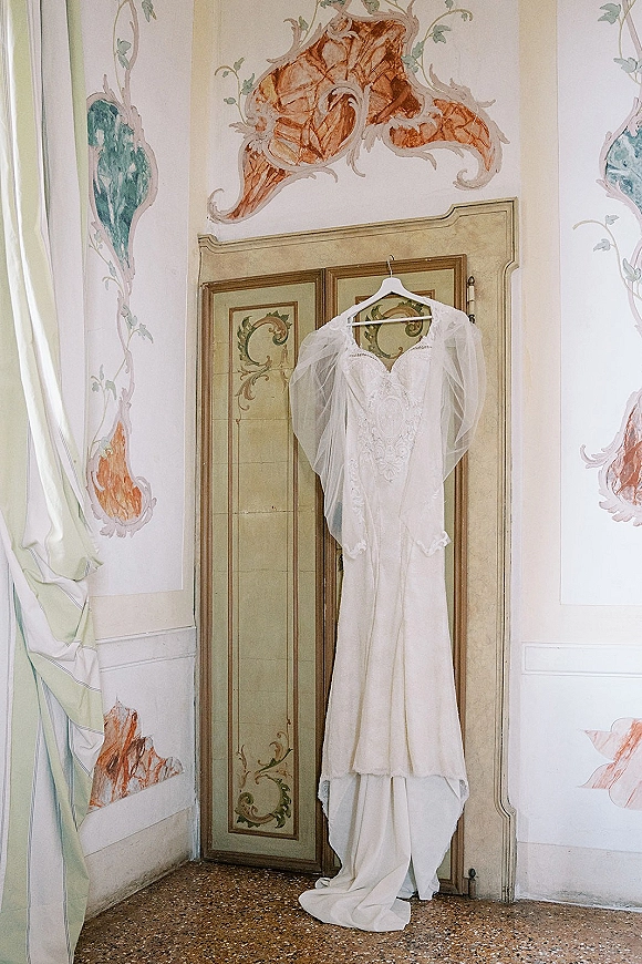 Wedding dress with lace bodice hanging on a hanger, framed by ornate double doors and painted wall murals, with tulle sleeves visible