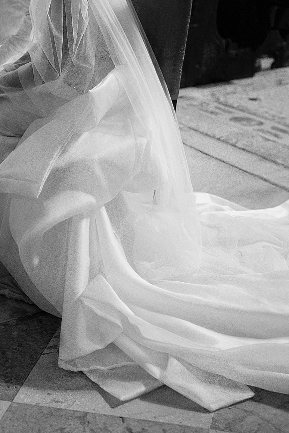 Wedding dress detail with a flowing wedding dress train and tulle veil draped over the hem on a stone floor in a shadowed interior