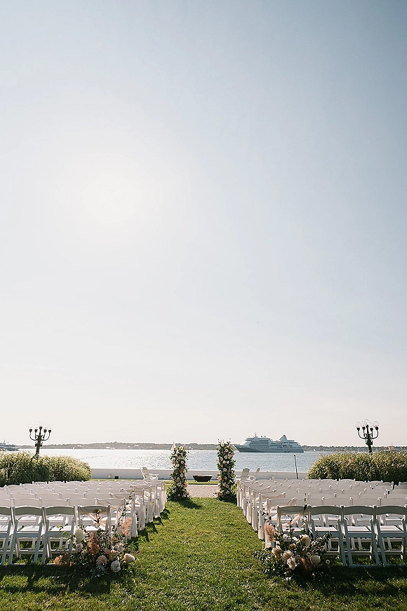 Ceremony setup with outdoor ceremony chairs lining a grass aisle, white and blush floral markers and pillars facing the ocean view coastline