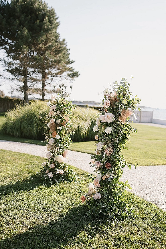 Wedding ceremony arch with an asymmetrical floral arch of blush and white roses and greenery, set on a coastal lawn with ocean view