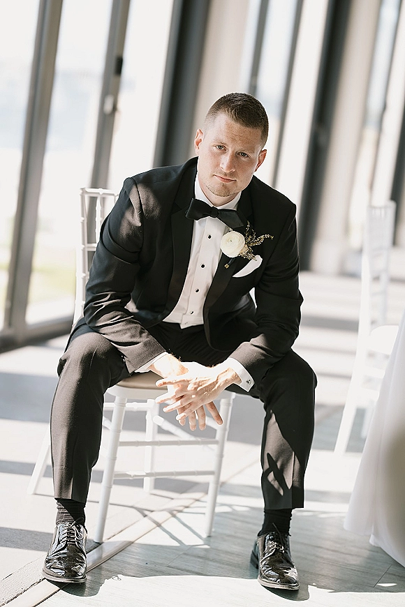 Groom portrait in a black tuxedo with white rose boutonniere, seated on a chair by large windows in natural light indoors