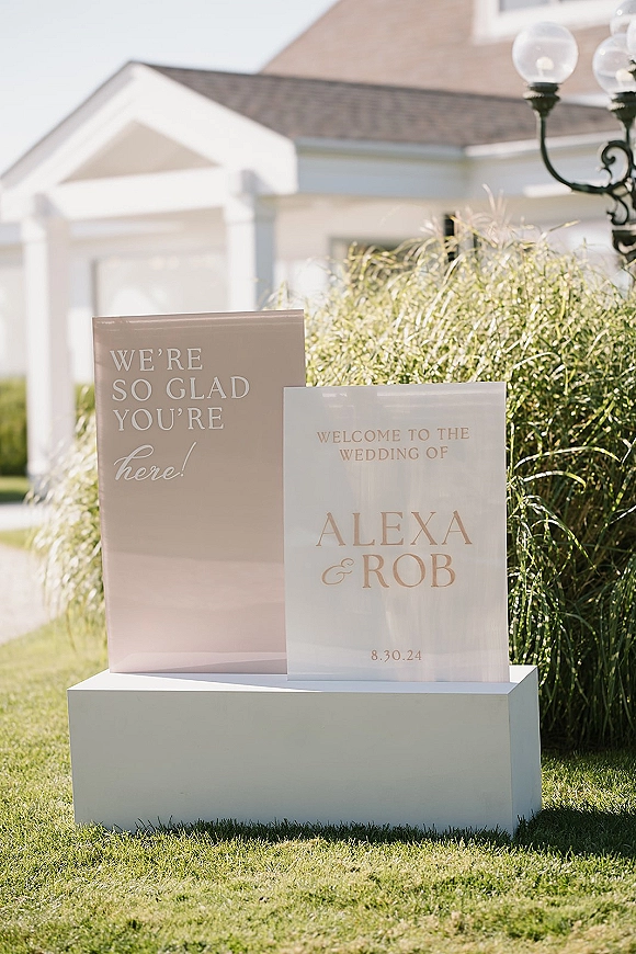 Wedding welcome sign in clear acrylic on a pedestal with ornamental grasses, set on a lawn outside a white building under blue sky