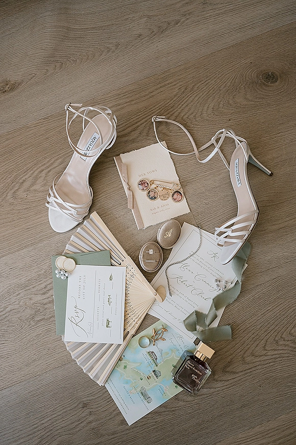 Wedding flat lay with wedding invitation suite, bridal heels, vow books, rings in a box, perfume bottle, ribbon, pins, and fan on wood floor
