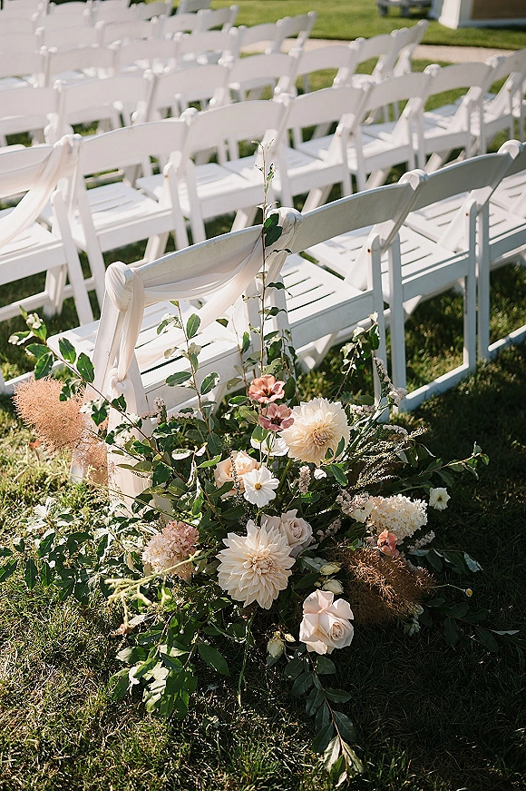 Ceremony aisle decor with outdoor ceremony seating, white folding chairs, draped fabric, and a low floral arrangement with greenery on a lawn