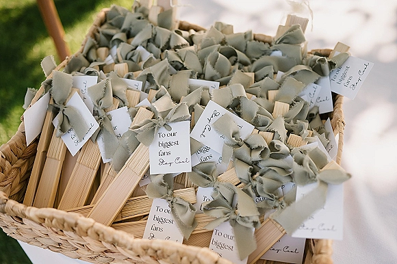 Wedding fan favors arranged in a woven basket, wooden handheld fan wedding favors with ribbon bows and calligraphy tags on green grass walkway