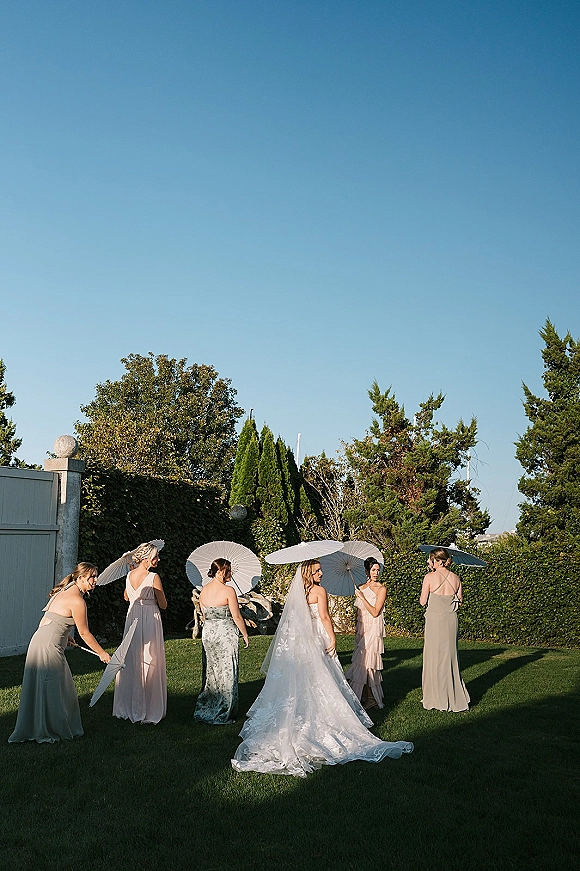 Bride with bridesmaids in a bridal party portrait, wearing a cathedral veil as bridesmaids hold white parasols in a sunny garden lawn
