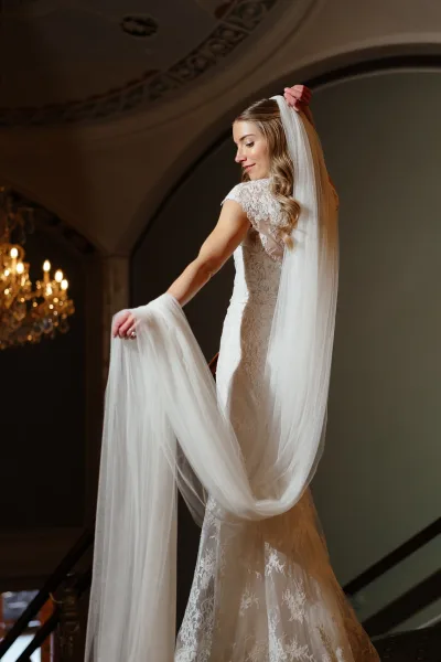 Bridal portrait of a bride holding veil in a lace wedding dress with long veil, posed on a grand staircase beneath a chandelier
