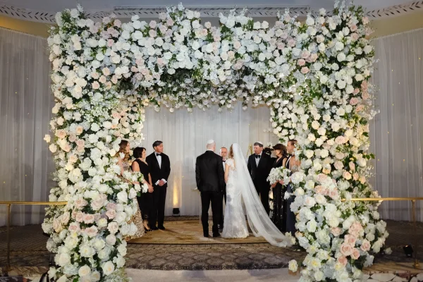Wedding ceremony setup with floral arch ceremony of white and blush roses, hydrangeas and draped fabric in an elegant indoor ballroom