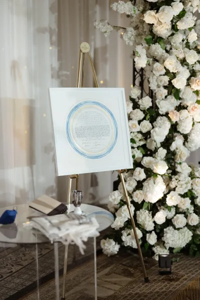Wedding ketubah display with framed artwork on a gold easel beside a ketubah signing table, set against a white rose floral wall with fairy lights