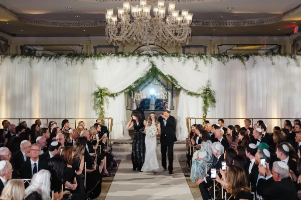 Wedding recessional as bride walking down aisle with bouquet beside groom in tux, beneath white drapery and chandelier in ballroom