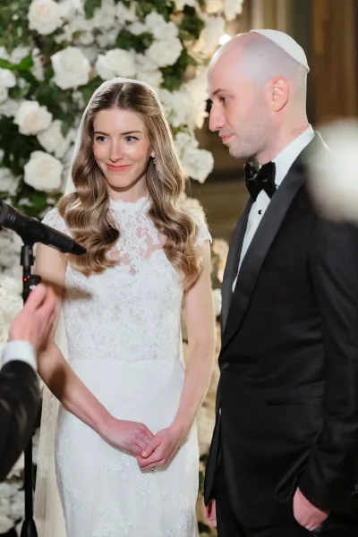 Ceremony moment during wedding vows as bride in lace dress and veil faces groom in tuxedo and kippah before a floral backdrop indoors