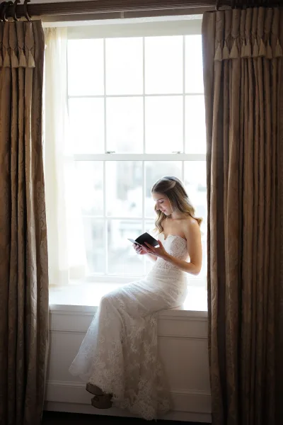 Bridal portrait of a bride sitting on a windowsill in a strapless lace gown, reading a book by a large window with curtains and soft light