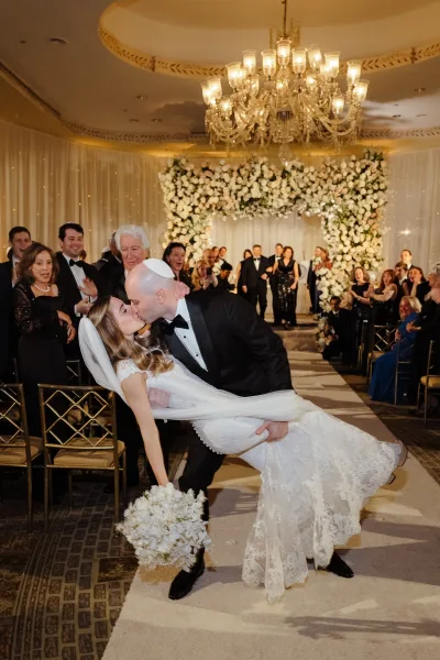 Wedding kiss as the groom dipping bride mid-aisle, her lace dress and veil flowing beneath chandeliers and string lights in a ballroom