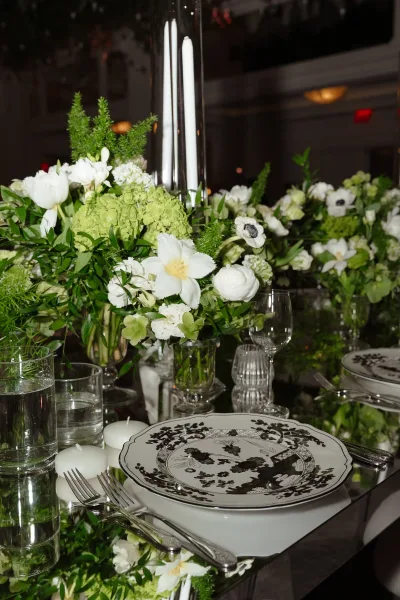 Reception tablescape with wedding table centerpiece of white and green florals, cylinder vases, taper candles, and black-and-white plates in a dim room