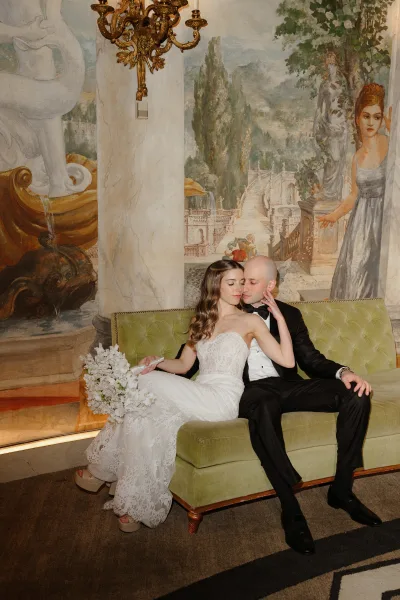 Couple portrait of bride and groom on sofa, cuddling on a green velvet couch beneath a gold chandelier against a mural wall backdrop