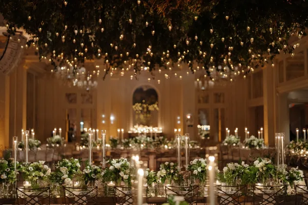 Reception tablescape with a candlelit wedding reception glow, white floral centerpieces, and hanging greenery under string lights in a grand ballroom