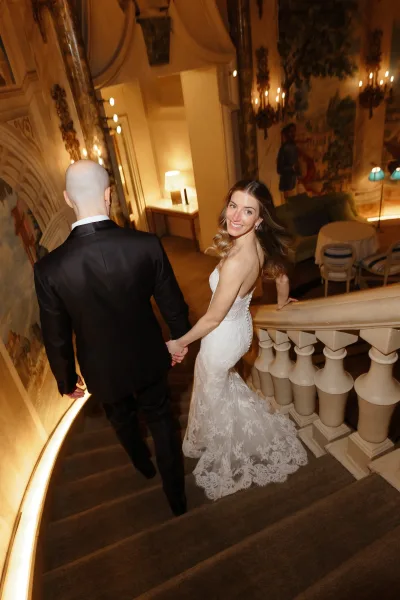 Couple portrait of bride and groom holding hands on a grand staircase, bride looking back in a lace gown with long train under warm lighting