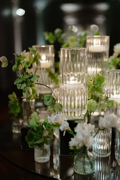 Wedding centerpiece with glass hurricane candle holders and pillar candles, white flowers and greenery on a mirrored table in a dim reception space