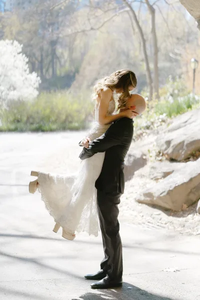 Wedding kiss portrait of groom lifting bride for a sunlit kiss on a park path, her lace wedding dress catching the backlight among trees and rocks