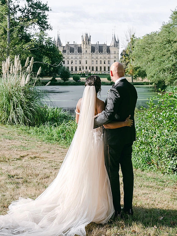 Couple portrait of bride and groom back view hugging, her long veil and gown train flowing, overlooking a lake with a castle beyond