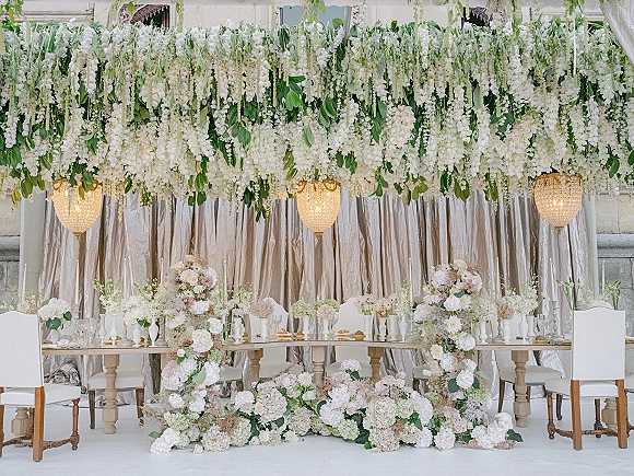 Reception sweetheart table with hanging floral installation, white blooms and greenery garland, candlelit centerpiece, crystal chandeliers, stone arch backdrop