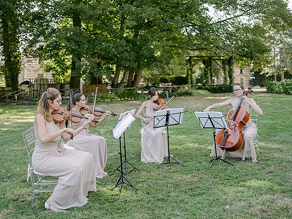 Wedding string quartet wedding ceremony musicians performing with sheet music on a garden lawn, with one blush dress among clear chairs
