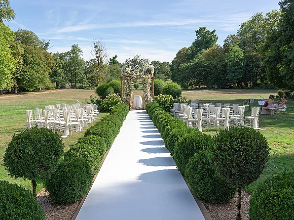 Outdoor ceremony setup with a floral arch, white aisle runner and clear acrylic chairs along a hedge-lined aisle on a sunny lawn