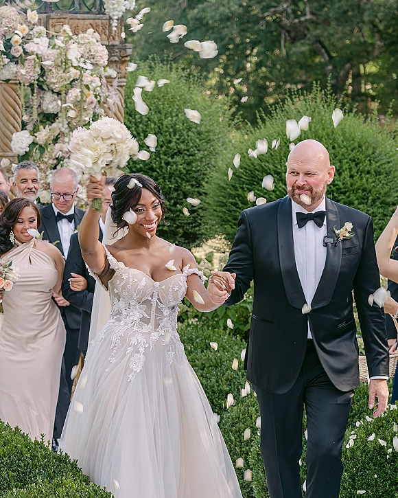 Wedding recessional as bride and groom walk hand in hand under a floral arch, bride raising bouquet while guests toss rose petals in a garden setting