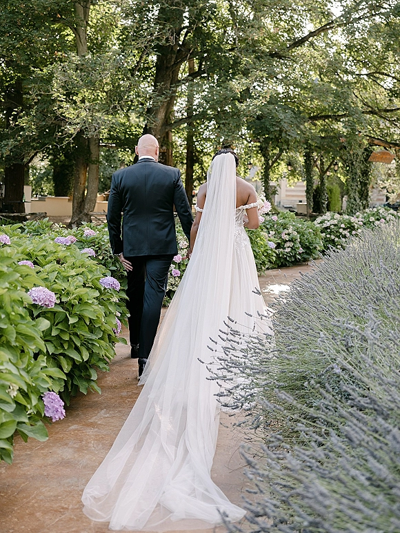 Couple portrait of bride and groom walking away hand in hand, her long veil trailing and bouquet in hand along a garden path