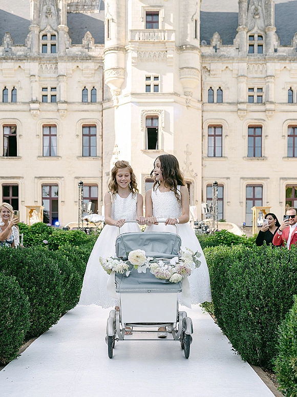 Flower girl processional with flower girls walking aisle, pushing a floral garland cart on a white runner past hedges and castle facade