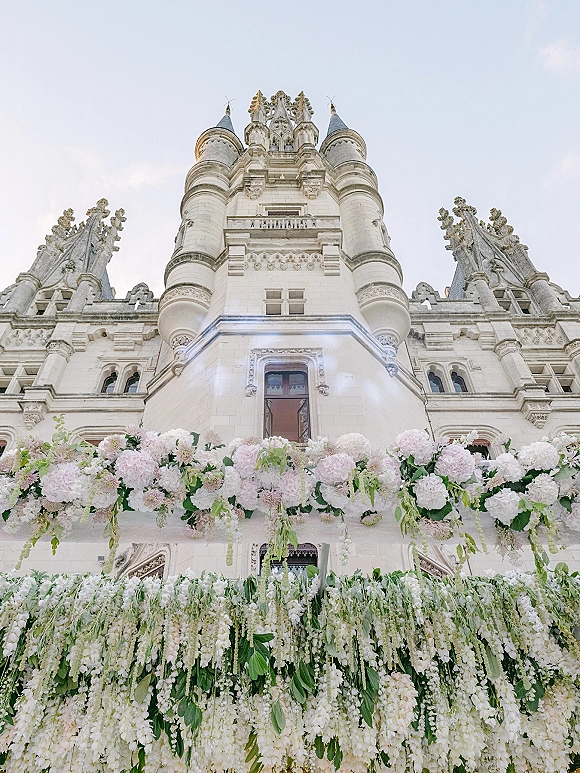 Wedding ceremony backdrop with cascading white and blush flowers and hanging greenery against a historic stone castle facade with towers