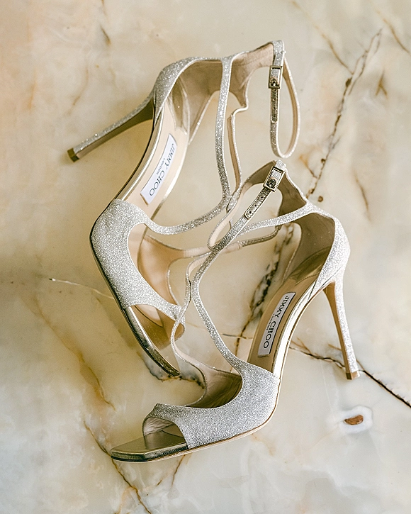 Bridal shoes, silver wedding heels with glitter finish, ankle straps and open toes, arranged in a flat lay on marble floor