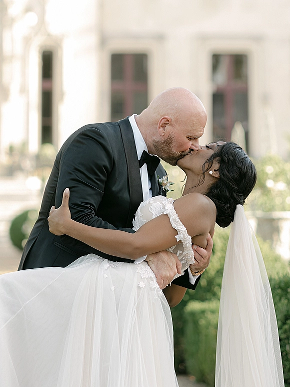 Wedding kiss portrait of groom dipping bride in lace gown and long veil, tuxedo and boutonniere, in a garden by stone estate windows