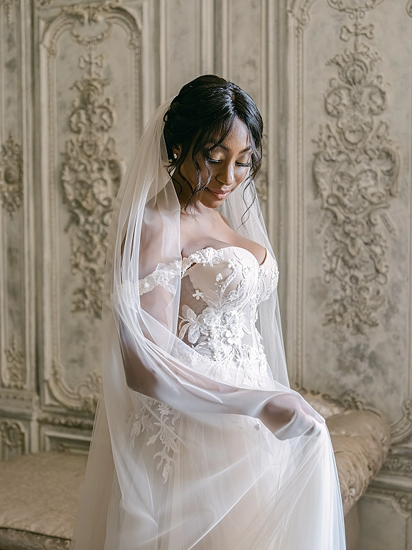 Bridal portrait of a bride in veil wearing a lace corset wedding dress, looking down beside ornate wall paneling in a vintage interior