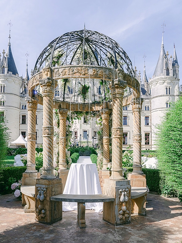 Wedding ceremony altar with white draped fabric on a wrought iron gazebo, floral garlands and hanging crystals in a castle garden setting