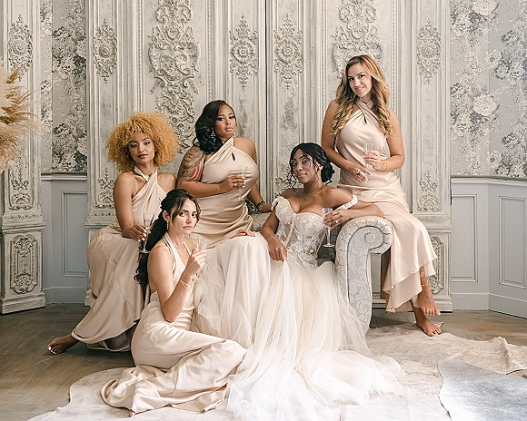 Bridesmaid group portrait with bride and bridesmaids in satin champagne dresses holding flutes before an ornate paneled wall backdrop