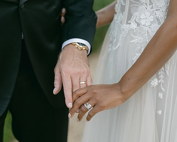 Wedding rings close-up on bride and groom hands, highlighting an emerald cut engagement ring and gold wedding band against greenery