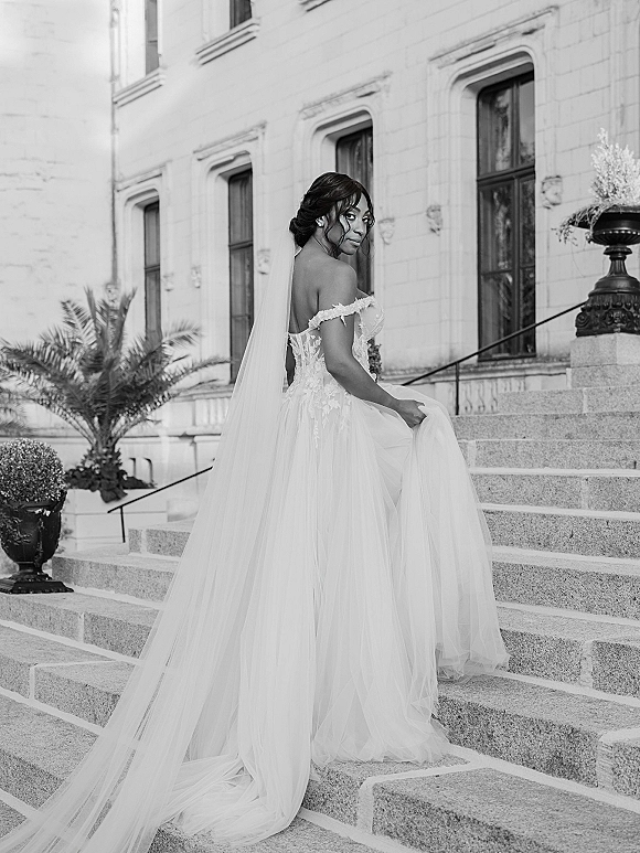 Bridal portrait of a bride on stairs in an off-the-shoulder lace gown, looking over her shoulder with a long veil on stone steps