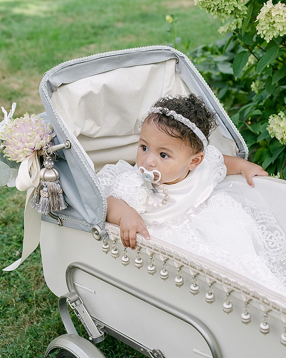 Flower girl portrait in a vintage baby carriage, wearing a white lace dress and floral headband with pacifier, on a green garden lawn