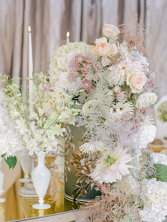 Wedding floral centerpiece with white and blush roses in a ceramic vase, mirrored table, and taper candles against neutral drapery backdrop