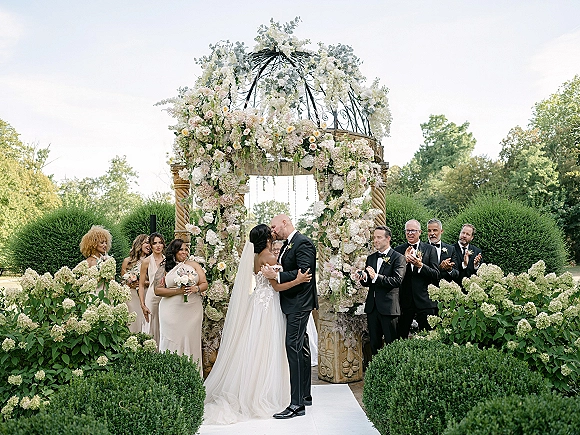 Ceremony kiss as bride and groom kiss under a floral arch, cathedral veil and tuxedo framed by a garden gazebo and hedges